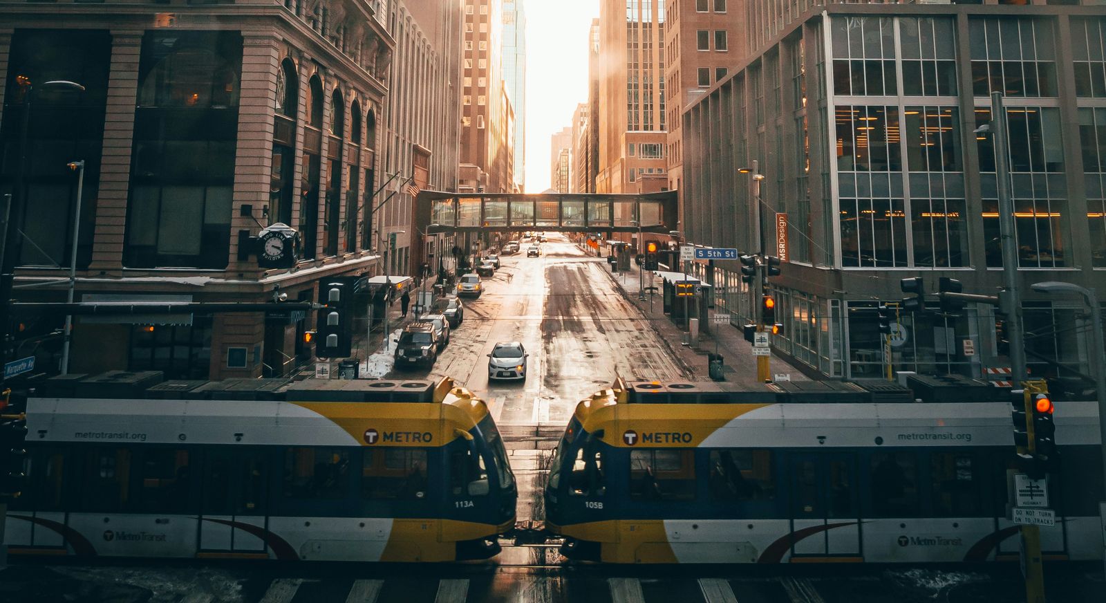 Two Metro Transit light rail trains meeting on 5th Street in downtown Minneapolis, with a skyway and sunlit office towers behind
